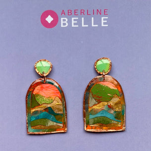 A pair of Aberline Belle handmade earrings displayed on a lavender card. The earrings feature a colorful, landscape-like mosaic design with green, orange, blue, and gold tones inside an arch-shaped frame, with a small green oval stone at the top.