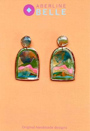 A pair of colorful handmade earrings with arch-shaped green and gold frames containing abstract landscape-like designs in pink, blue, and green, displayed on a peach background with the brand name Aberline Belle.