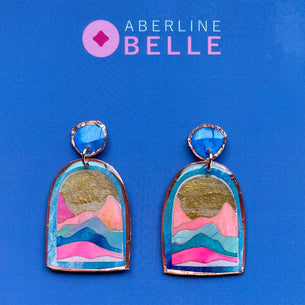 A pair of colorful handmade earrings displayed on a blue card. The earrings feature a blue oval top and an arch-shaped pendant with a hand-painted mountain landscape design in pink, blue, teal, and gold leaf.