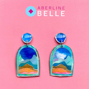 A pair of Aberline Belle handmade earrings displayed on a solid pink background. The earrings feature a colorful landscape design with blue mountains, green hills, and orange sand, suspended from round blue beads.