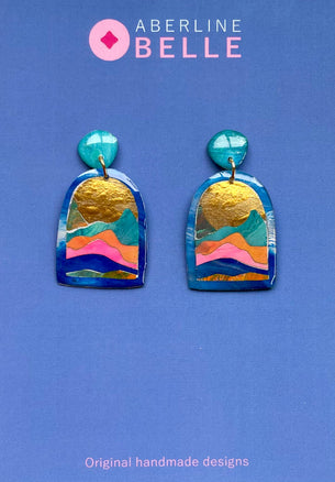A pair of handmade earrings by Aberline Belle, featuring a blue arched pendant with a colorful layered landscape design and textured gold leaf accents, displayed on a solid blue background.