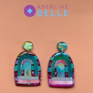 A pair of handmade arch-shaped earrings with colorful mosaic details in turquoise, green, pink, and purple, displayed on a peach background with the brand name 'Aberline Belle' at the top.