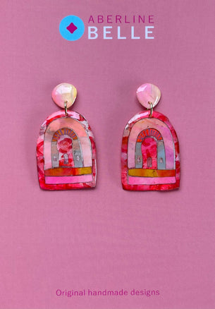 A pair of pink and red arch-shaped acrylic earrings with a colorful printed design, displayed on a solid pink background with the Aberline Belle brand logo at the top.