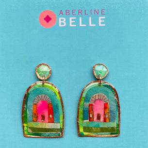 A pair of colorful statement earrings by Aberline Belle, featuring a hand-painted archway design in green, pink, and gold, displayed on a light blue background.