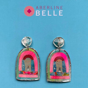 A pair of handmade arch-shaped earrings with silver-toned metal borders, bright pink inner arches, and multicolored collage details, displayed on a solid blue background with the brand name 'Aberline Belle' at the top.
